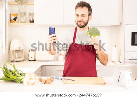 A man is taking a picture of a broccoli in his kitchen. He is wearing a red apron and standing in front of a counter with a knife and a cutting board