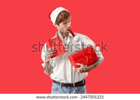 Surprised young man in Santa hat with gift card and Christmas present on red background