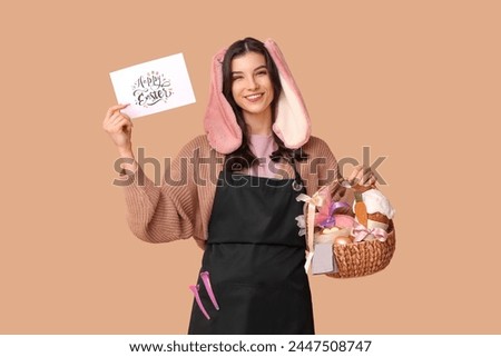 Happy female hairdresser in bunny ears with greeting card and Easter basket on brown background
