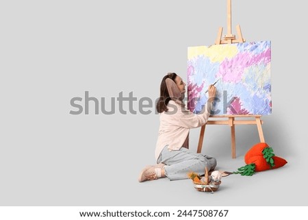 Happy female artist in bunny ears with Easter basket, toy carrots painting picture on grey background