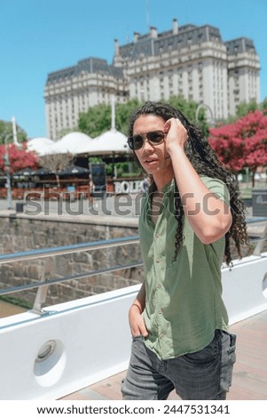 Vertical image of young Latin man with curls in green clothes and sunglasses on vacation in Argentina walking through city at noon, image for vacation social networks.