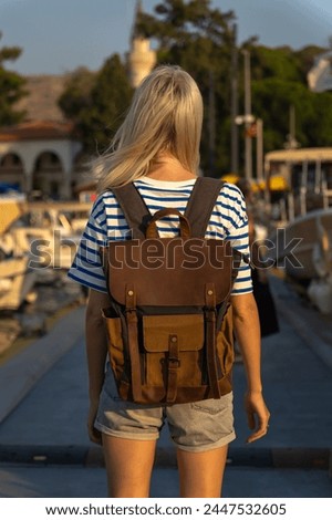 Young female tourist with a craft backpack stands on the pier near yachts and boats at sunset, back photo, Marmaris, Turkey. Concept of travel and tourism