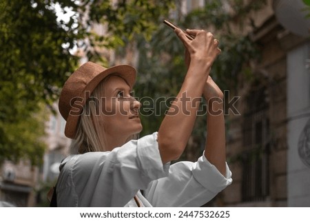 Stylish young blonde woman in a hat, white shirt with a backpack takes pictures on a beautiful street in the city on a mobile phone. Young female traveler