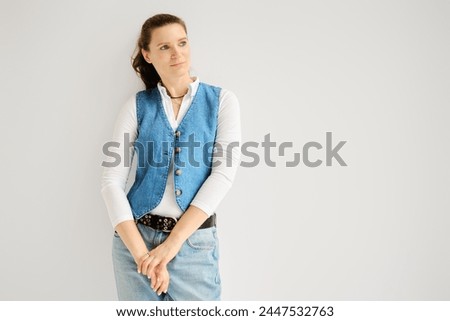 Portrait of relaxed woman in denim vest, jeans and cotton shirt leaning against white wall