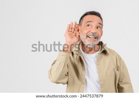 Elderly man with a beard hand to ear gesture indicating listening intently or hearing difficulty on light background