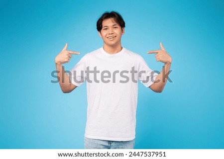 Self-assured teen asian guy in a white shirt pointing at himself against a solid blue studio background Royalty-Free Stock Photo #2447537951