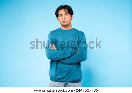 Asian young guy stands confidently with arms crossed against a blue background, showcasing attitude and style Royalty-Free Stock Photo #2447537985