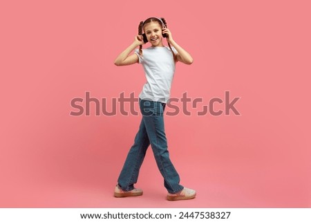 A teen girl casually walks while enjoying music from her headphones on a pink studio background, full length