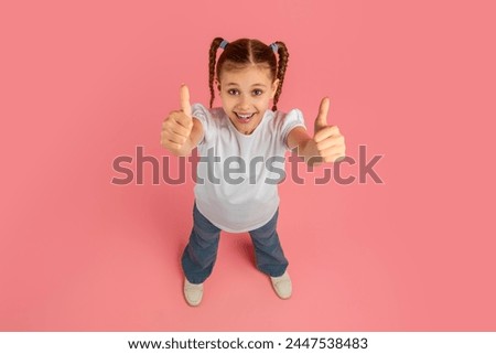 Confident and happy girl looks at the camera, giving two thumbs up against a pink background, signaling approval Royalty-Free Stock Photo #2447538483