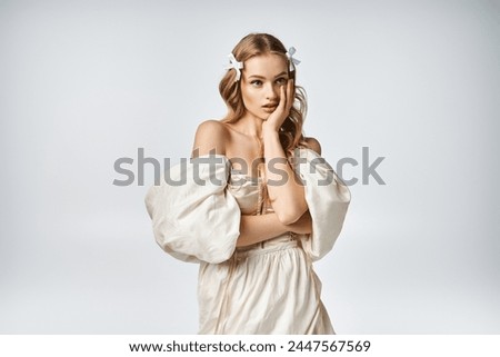 A young blonde woman exudes elegance in a studio setting, wearing a white dress while striking a pose for a picture.