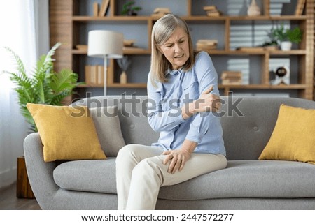 Mature woman having elbow injury and massaging sore spot with hand in stylish apartment interior. Unhealthy female in light clothes having tension in muscles while looking aside on hurting area. Royalty-Free Stock Photo #2447572277