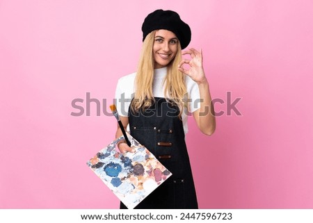 Young artist woman holding a palette over isolated pink background showing an ok sign with fingers