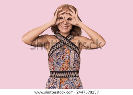Redhead mid-aged Caucasian woman in studio showing okay sign over eyes