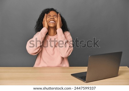 African American at desk with laptop laughs joyfully keeping hands on head. Happiness concept.