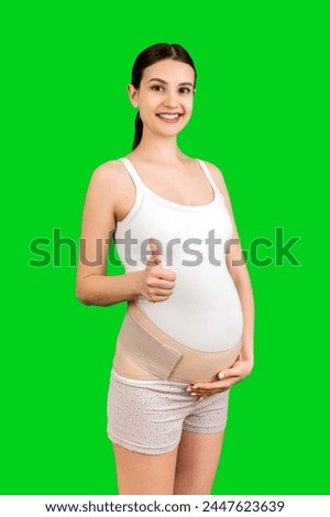 Portrait of young pregnant woman wearing bandage on her belly and showing thumb up sign at green background with copy space. Orthopedic abdominal support belt concept.