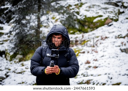 Frontal photo captures young adult in black coat and hood, recording snowy woodland scene. Royalty-Free Stock Photo #2447661593