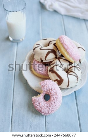 Frosted vanilla donuts with chocolate swilrs and strawberry pink doughnuts with coconut flakes. Bite missing. Glass of milk in background. Selective focus with blurred foreground and background.