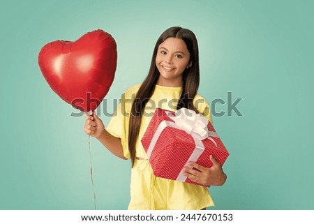 Teenager child holding gift box on blue isolated background. Gift for kids birthday. Christmas or New Year present box. Happy girl face, positive and smiling emotions. Love valentines, heart balloon.