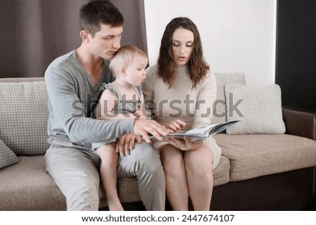 Child sits in his dad arms, his mom reads a book to toddler, looking at educational pictures. Parents spend time with their baby together, sitting on couch at home. Concept of family, child learning