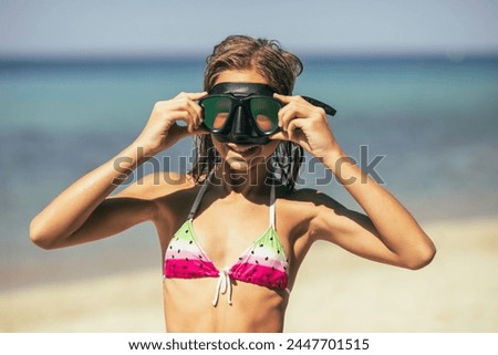 A joyful teen girl in a bikini and diving mask standing on the sandy beach, ready for a swim or dive in the ocean waves on a sunny day.