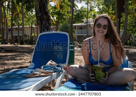 A woman sits on a lawn chair on the beach with a coconut in her hand at a tropical tourist resort