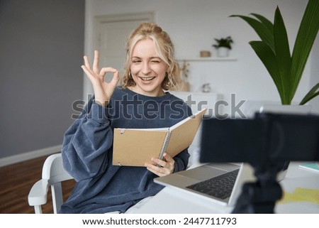 Portrait of cute blond girl with notebook in hands, shows okay sign, records video, social media content, blogging from her room.