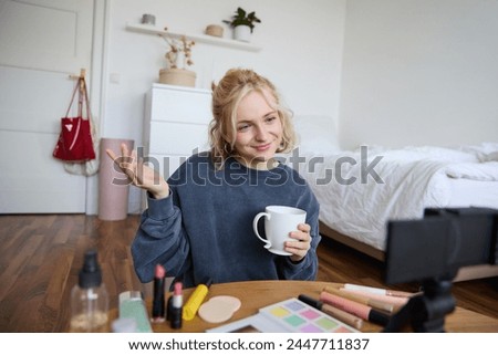 Image of young woman, makeup vlogger, sitting in bedroom with digital camera, drinking tea and talking, creating lifestyle video, social media content.