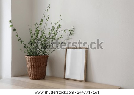 Spring, Easter still life. Elegant Scandinavian living room, home office. Empty vertical wooden picture frame mockup on desk, table. Wicker willow basket with green birch tree branches, side view.