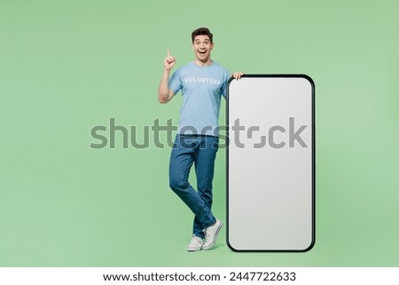 Full body young man wear blue t-shirt title volunteer big huge blank screen area mobile cell phone point finger up isolated on plain green background. Voluntary free work help charity grace concept