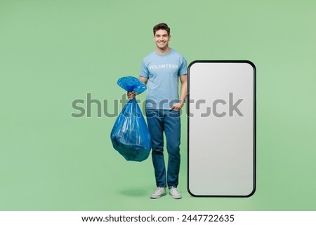 Full body young man wears blue t-shirt title volunteer big huge blank screen area mobile cell phone hold trash bag isolated on plain green background. Voluntary free work help charity grace concept