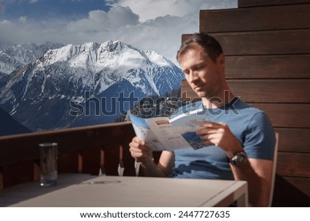 Man reading brochure outdoors at wooden table with water glass, mountains in background. Relaxed setting suggests mountain resort or scenic viewpoint. Royalty-Free Stock Photo #2447727635