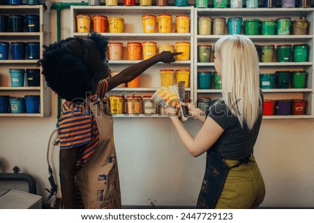Back view of multicultural graphic industry female workers standing near shelves with pain buckets and choosing colors on color swatch in printing shop. Female interracial entrepreneurs in workshop.