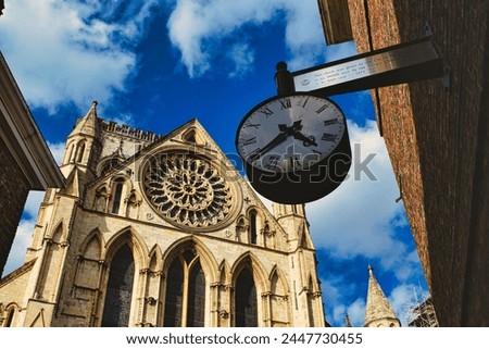 Vintage street clock hanging with a gothic cathedral facade in the background, showcasing intricate architecture and a clear blue sky in York, North Yorkshire, England.