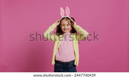 Adorable cute child putting bunny ears and waving at camera, enjoying easter sunday celebration against pink background. Smiling cheery schoolgirl with pigtails saying hello. Camera B. Royalty-Free Stock Photo #2447740439