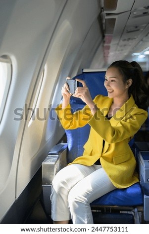 A woman in a yellow jacket is taking a picture of the view from an airplane window. She is smiling and she is enjoying the experience