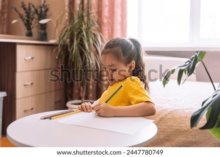 A child draws at the table with a green pencil on a sheet of paper.