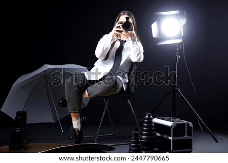 Female photographer with modern camera and lighting equipment taking picture of viewer on black background