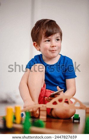 A joyful young boy with a smartphone sits amidst a wooden train setup, his bright laughter echoing the playful fusion of digital and tangible play Royalty-Free Stock Photo #2447827659