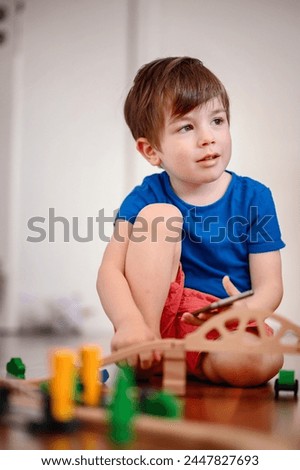 A joyful young boy with a smartphone sits amidst a wooden train setup, his bright laughter echoing the playful fusion of digital and tangible play Royalty-Free Stock Photo #2447827693