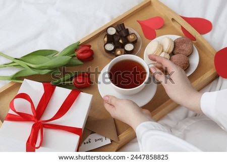 Tasty breakfast served in bed. Woman with tea, desserts, gift box and flowers at home, closeup