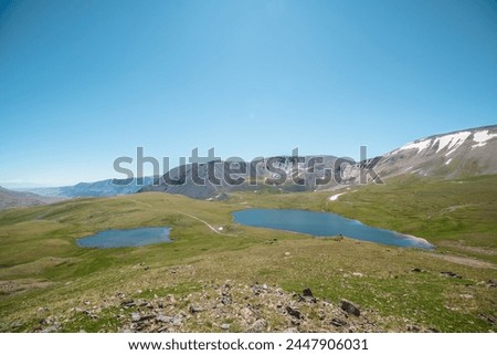 Two lakes, sunlit green meadow, alpine valley and mountain range. Blue sky gradient reflected in water. Top view from rocks to dirt road between two beautiful lakes in high mountains in sunny day. Royalty-Free Stock Photo #2447906031