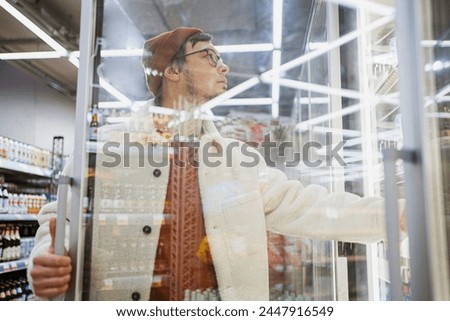 A contemplative shopper in a warm jacket and beanie examines products through the glass doors of a refrigerated section in a well-lit grocery store. Man in Winter Attire Browsing Products in Royalty-Free Stock Photo #2447916549
