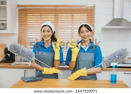 Two Asian young cleaning service women on kitchen counter with duster foggy spray and rag showcase efficient housework teamwork. Clean portrait two uniform maid working smiling employee. Royalty-Free Stock Photo #2447920071