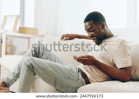 African American Man Sitting on Black Sofa, Using Smartphone and Typing Message at Home Modern Technology and Relaxation Concept This image depicts a young African American man enjoying his leisure
