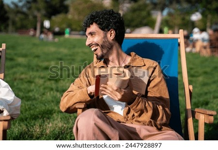 Side view of smiling young bearded Hispanic male looking away while sitting with book on deckchair and enjoying joke of unrecognizable friend relaxing on green park against blurred trees Royalty-Free Stock Photo #2447928897