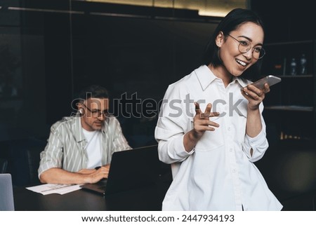 Cheerful female in classic glasses for eyes correcting connecting to 4g wireless on cellular gadget for sending audio sms message during online chatting, happy Asian woman recording sound