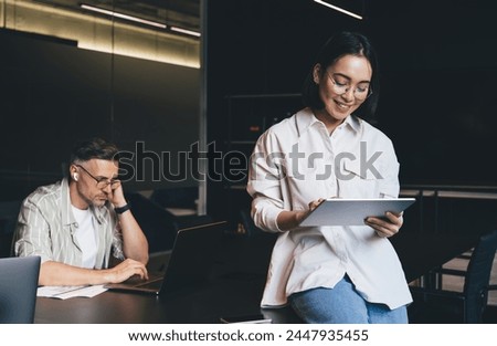 Happy Asian employee in classic eyewear for vision correction using digital tablet for browsing information on website via application, successful graphic designer in spectacles making online booking