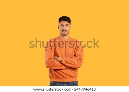 A stern-looking man with moustache stands with arms crossed on a solid yellow background, copy space Royalty-Free Stock Photo #2447946413