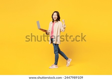 Full body young IT woman of Asian ethnicity she wear pink t-shirt beige shirt pastel casual clothes hold use work on laptop pc computer isolated on plain yellow background studio. Lifestyle portrait