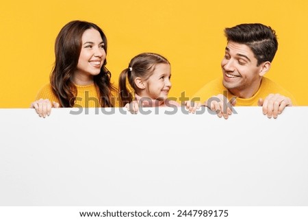 Young parents mom dad with child kid girl 7-8 years old wear pink casual clothes hold big blank poster billboard for content look to each other isolated on plain yellow background. Family day concept